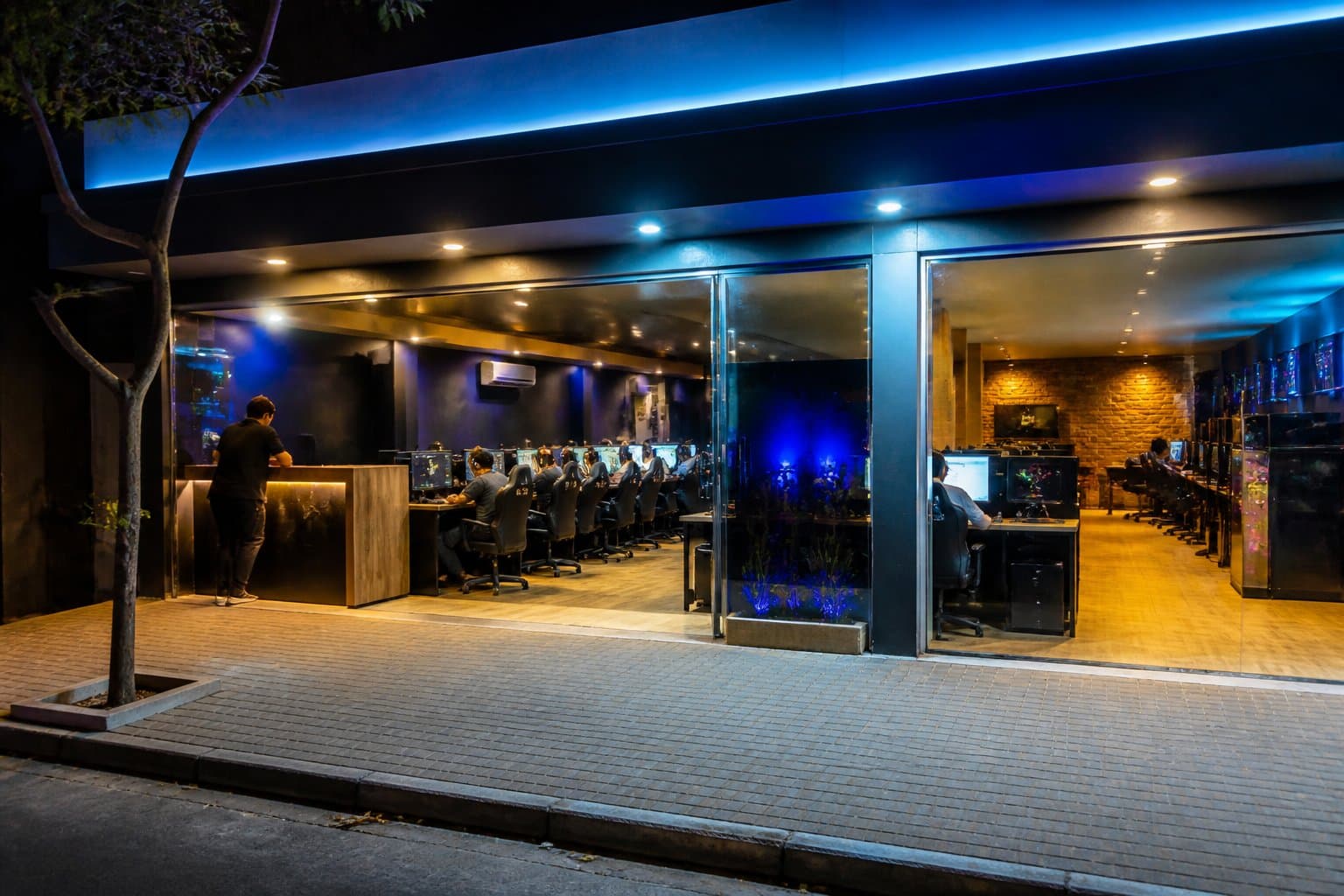 Lan house iluminada e aberta à noite, com computadores e pessoas usando as estações no interior.